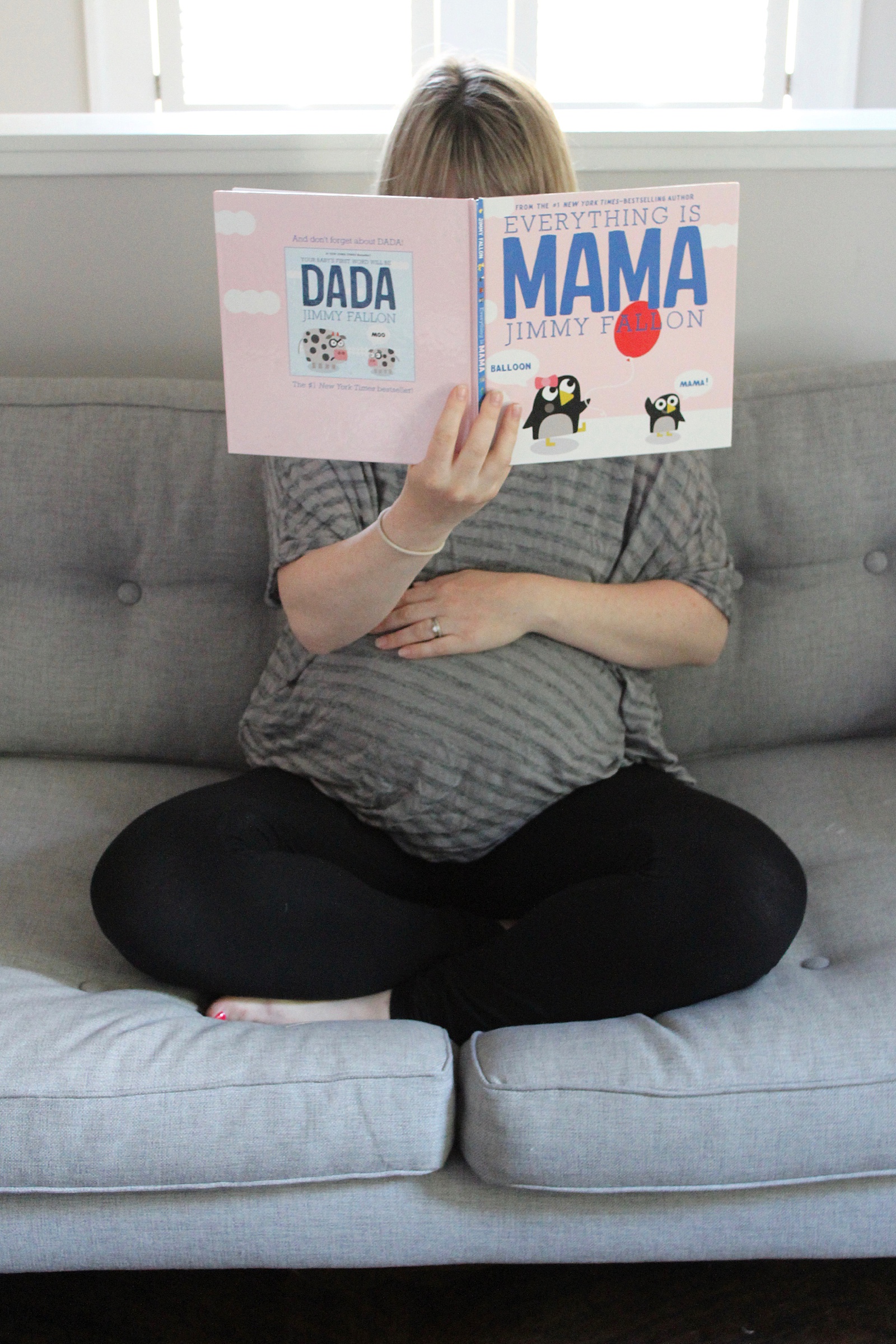 A pregnant woman sits on a couch reading a children's book.