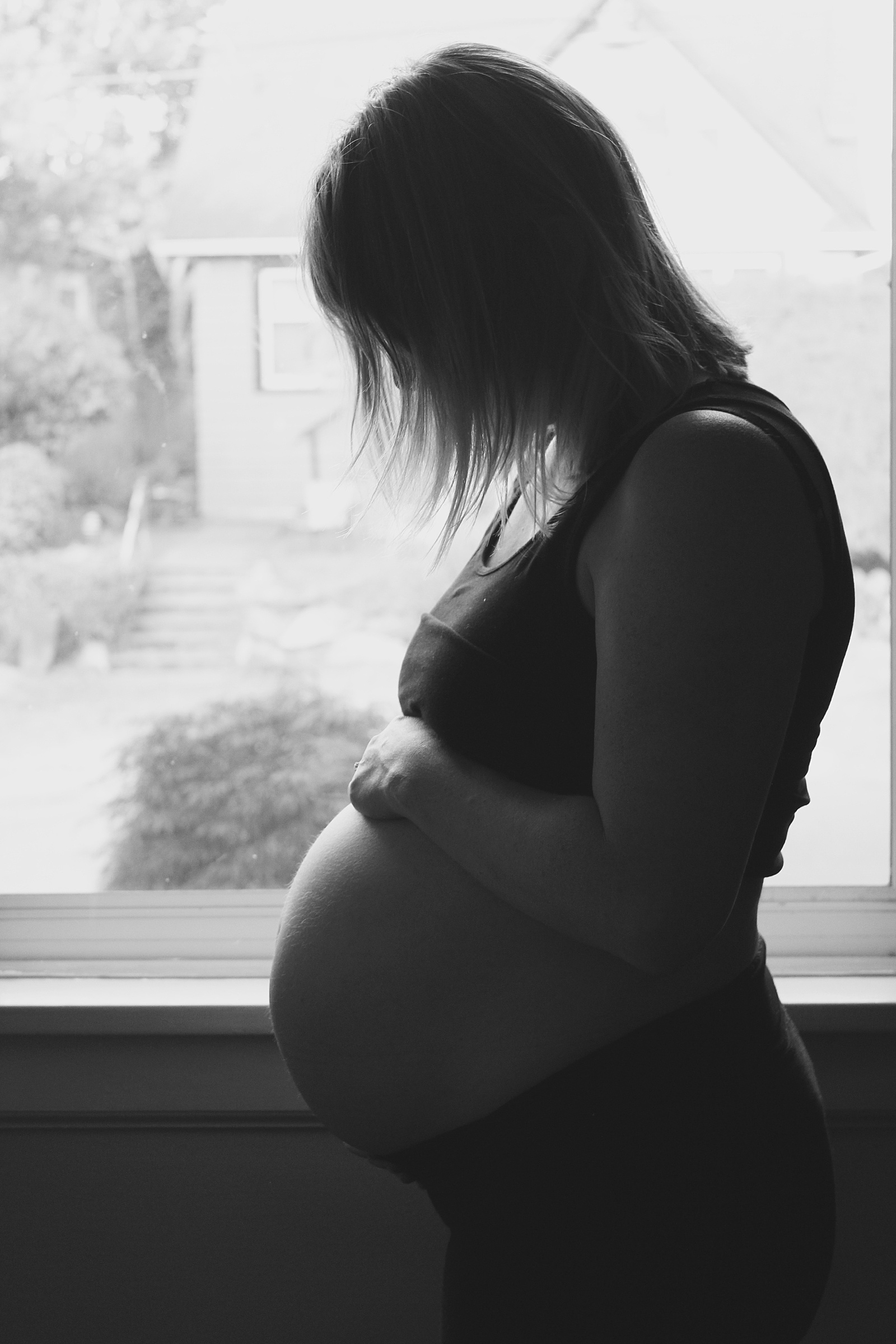 A woman stands backlit by a window for a baby bump pic.