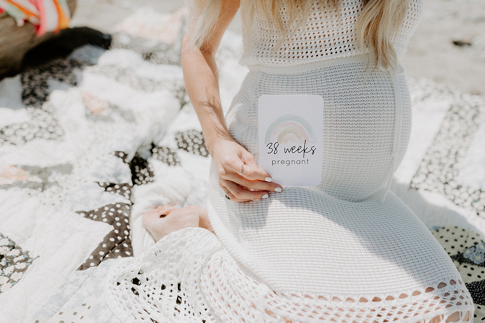 A pregnant woman holds up a pregnancy moment card in a maternity photo on a crocheted blanket.