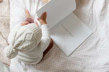 A toddler opens a baby book sitting on a bed.