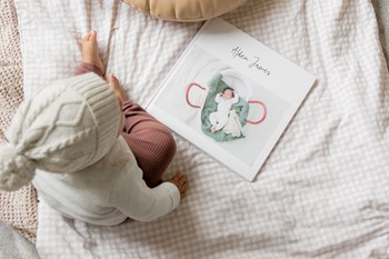 A toddler sits on a bed and poses with a custom baby book.